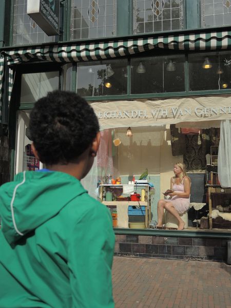 Een jongen in een groene jas kijkt naar de etalage waarin een vrouw in een roze jurk zit te lezen, met een groene papegaai naast zich.