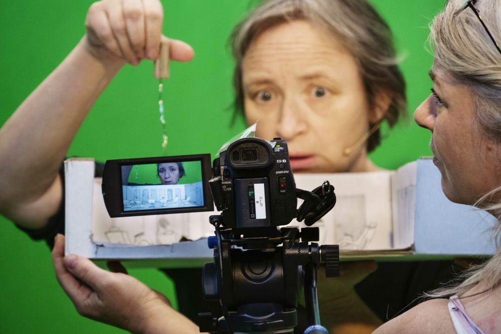 Twee speelsters houden een kartonnen decorstuk bij de camera; de een laat er een poppetje in zakken, op het cameraschermpje verschijnt een gezicht.