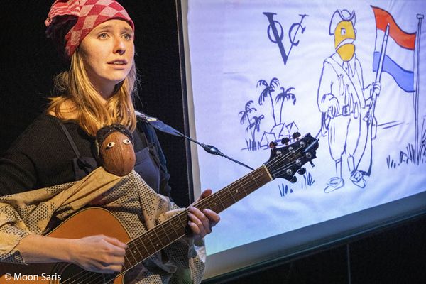 Een speelster met een rood geblokte hoofddoek speelt gitaar met een pop op schoot; achter haar een tekening met het VOC-logo, een soldaat en de Nederlandse vlag.