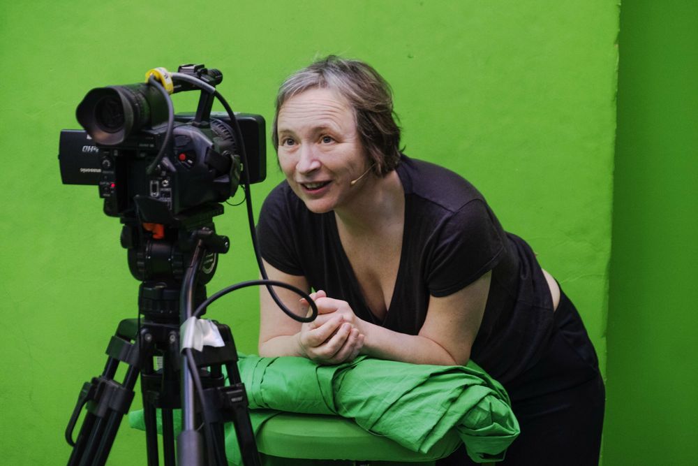 Een speelster leunt lachend met een microfoontje op naast de camera in de groene studio.