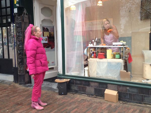 Een vrouw in een felroze jas kijkt van de stoep naar de speelster die in de etalage aan haar tafeltje staat.