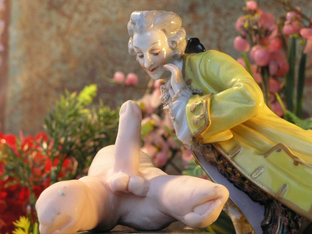 Porseleinen beeldje: een heer in een geel rococojasje buigt zich over een naakt figuurtje dat voor hem ligt, tussen roze bloemen.