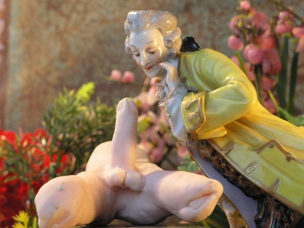 Porseleinen beeldje: een heer in een geel rococojasje buigt zich over een naakt figuurtje dat voor hem ligt, tussen roze bloemen.