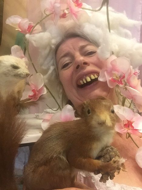 Zelfportret van dichtbij: een speelster met een pruik van watten en orchideeën lacht met opgezette eekhoorns in haar armen.