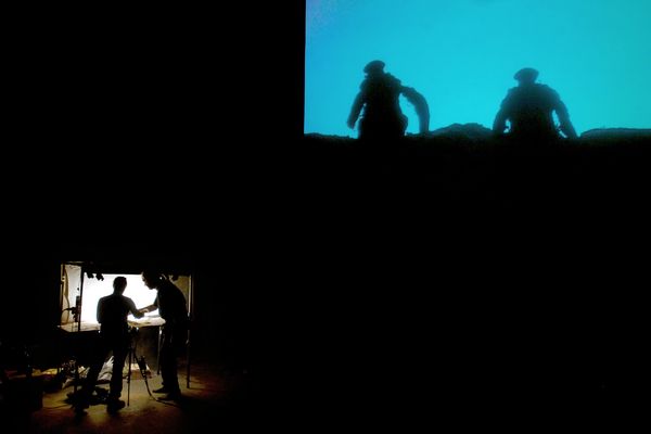 Toneelbeeld: op het scherm de silhouetten van twee soldaten tegen een blauwe lucht, daaronder filmen twee makers in een verlicht nisje de maquette.