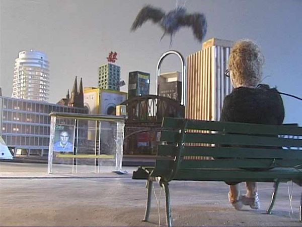 Een oude vrouw zit op een bankje voor de skyline van een miniatuurstad; boven haar vliegt een vogel voorbij.