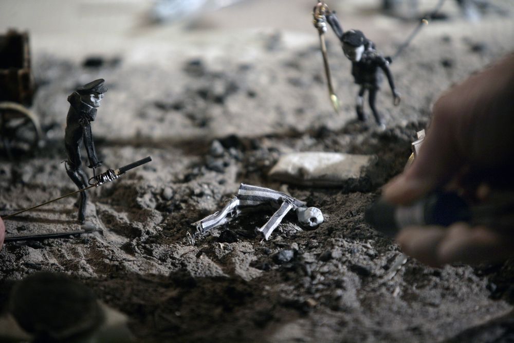 In de maquette ligt een gevallen poppetje op de grond tussen twee bewakersfiguren; rechts komt de hand van een maker in beeld.