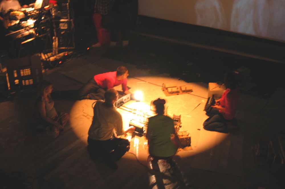 Vijf spelers zitten in een lichtcirkel op de toneelvloer gebogen over het kleine decor, met camera's en kabels om zich heen.