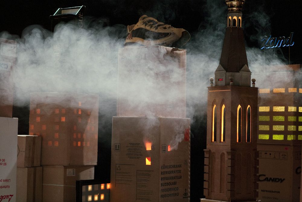 Skyline van kartonnen dozen in de rook: op een van de torens staat een sportschoen, ernaast een kerktoren met verlichte ramen en een blauw neonbord.