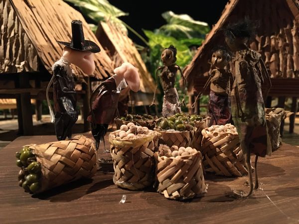 Een pop met hoge hoed staat tussen gevlochten manden vol noten, met palmbladhutten en dorpsbewoners op de achtergrond.