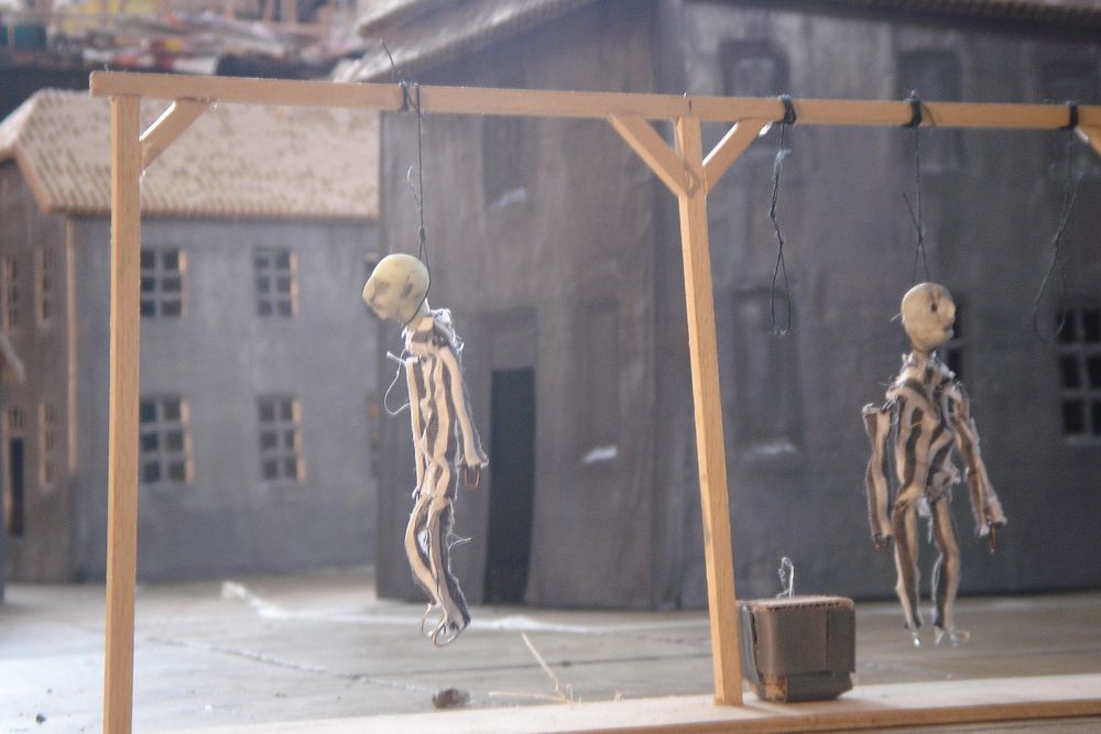 Een galg met twee opgehangen poppetjes staat in de maquette voor een grijs gebouw.