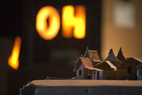 Het oranje lichtbord OH gloeit onscherp achter een dorpje van kartonnen huisjes met puntdaken.