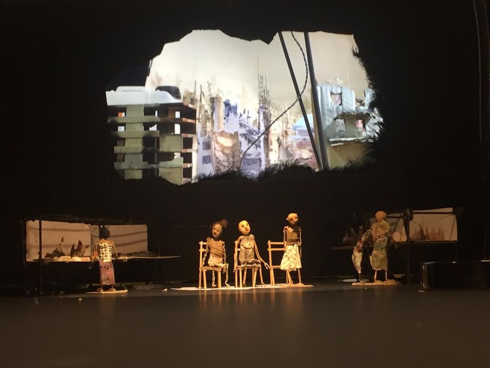 Toneelbeeld: poppen zitten op stoeltjes op het lege podium, daarboven de projectie van een verwoeste stad in een gescheurde vorm.