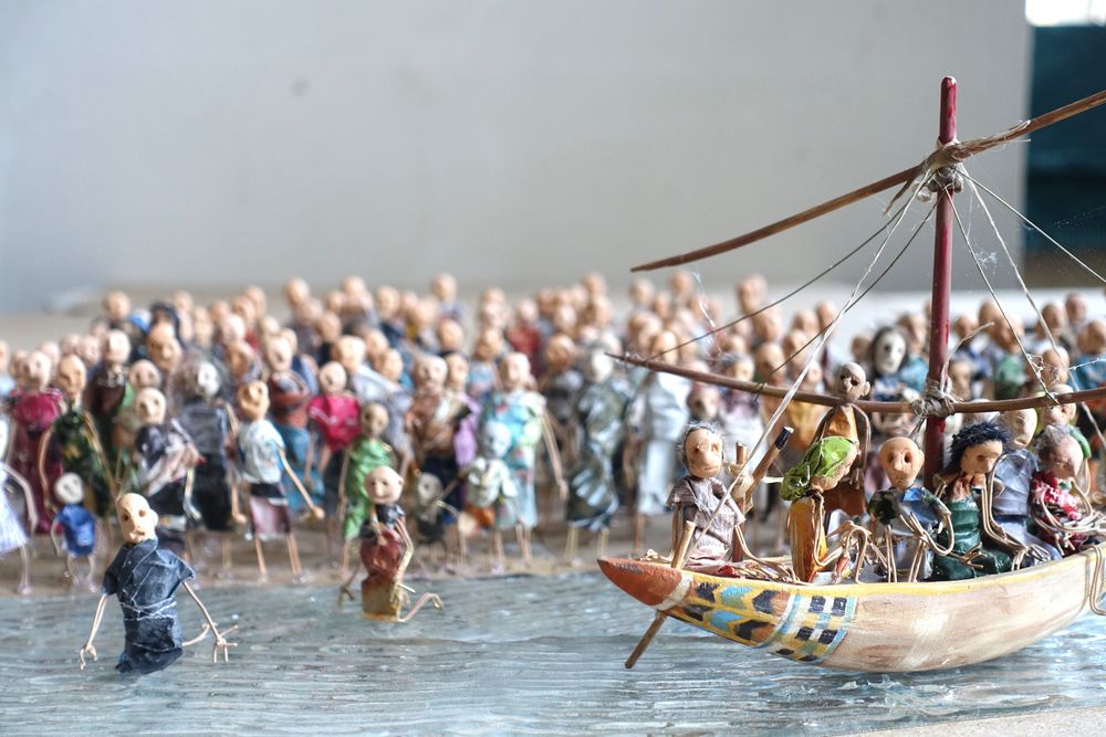 Een menigte poppetjes waadt door het water naast een volgeladen boot met een gepunte mast.