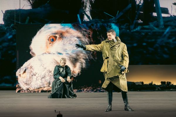 Op het scherm de reusachtige kop van een vogel; ervoor knielt een speelster en wijst een speler in een groene jas met uitgestrekte arm.