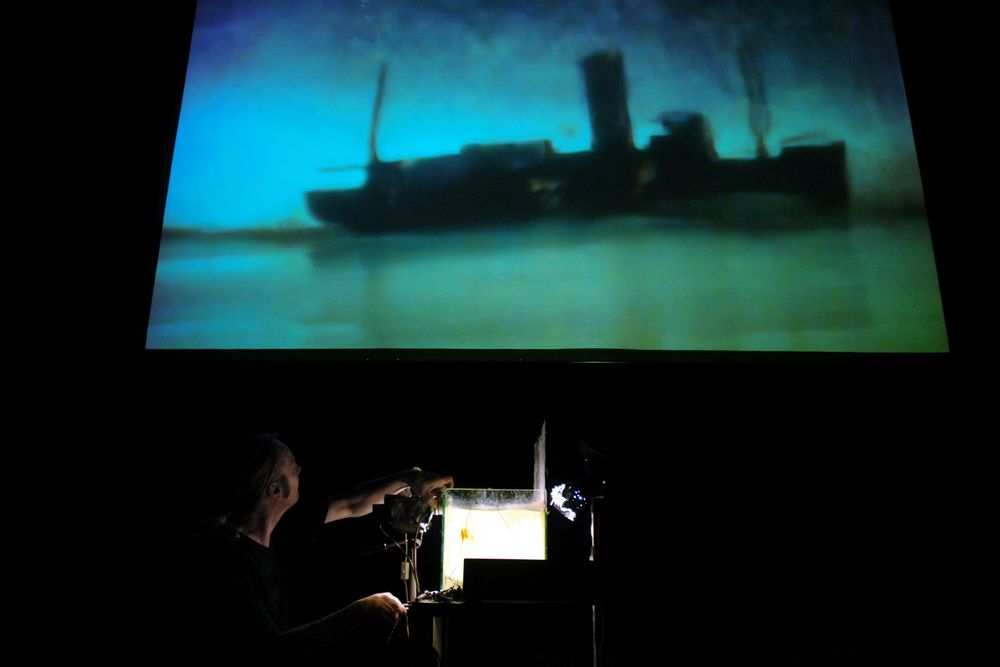Toneelbeeld: op het scherm een schip in blauwgroene nevel, daaronder filmt een maker het kleine decor met een camera.