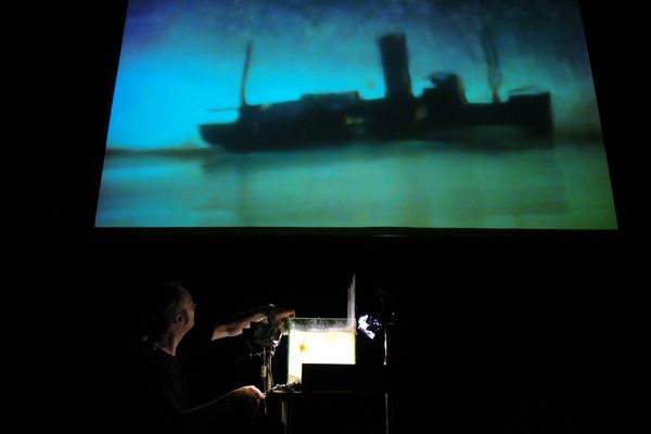 Toneelbeeld: op het scherm een schip in blauwgroene nevel, daaronder filmt een maker het kleine decor met een camera.