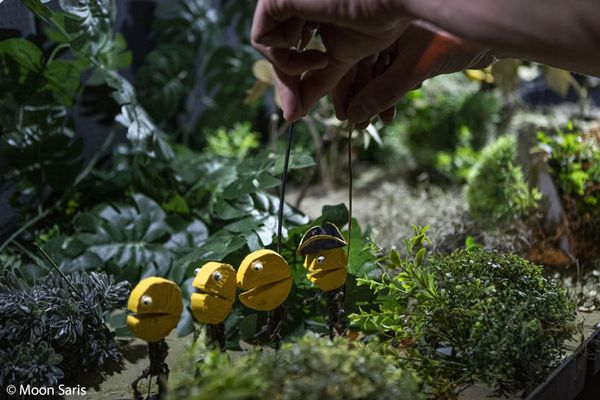 Een hand zet aan draadjes vier gele poppetjes met een grote mond neer in een decor van echte plantjes.