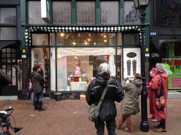 Straatbeeld: een groepje mensen kijkt vanaf de stoep naar de vrouw in de verlichte etalage van de boekhandel.