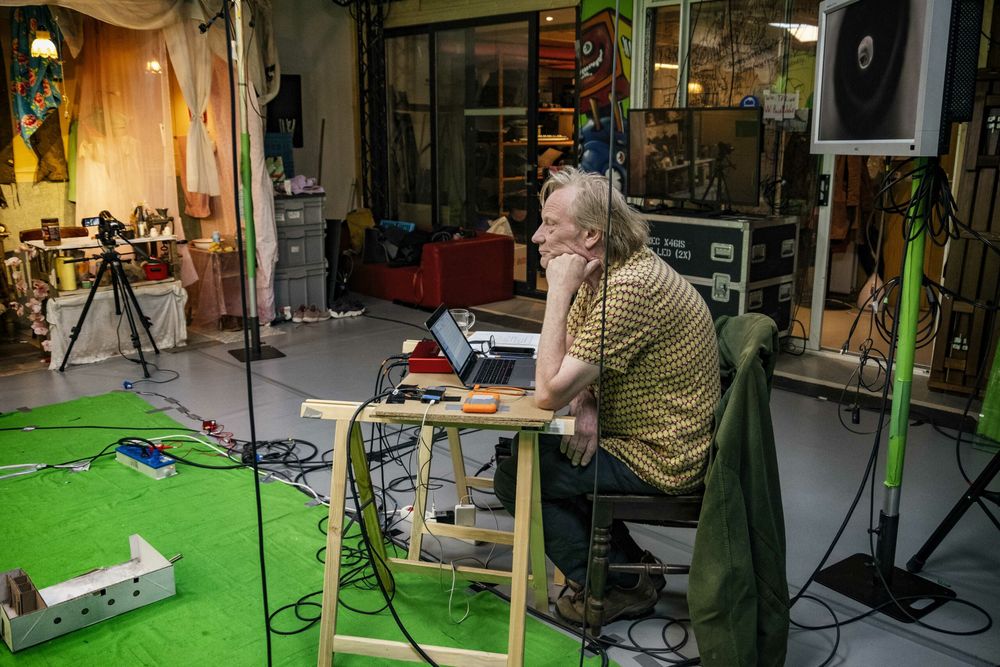 Een maker zit met zijn hoofd in zijn hand aan een laptop in de studio, tussen groene doeken, statieven en kabels.