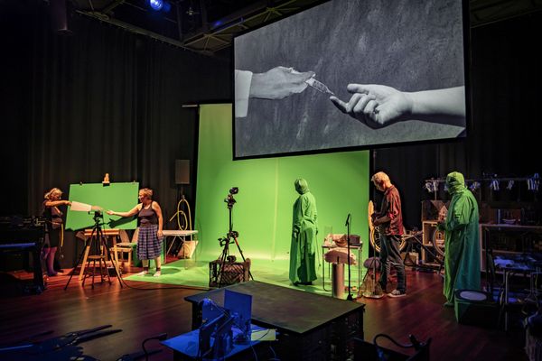 Wijd toneelbeeld van de green screen-studio: spelers en camera's rond een groen doek, één speler in een groen pak, met daarboven het geprojecteerde beeld van twee handen.