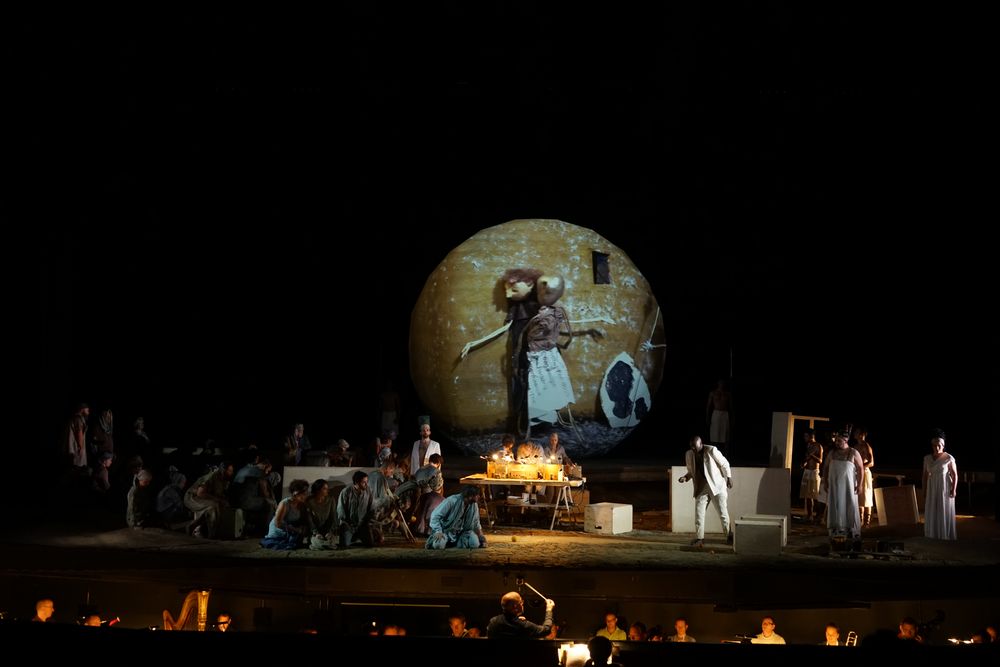 Wijd beeld van de opera met de grote bol waarop een figuur met een staf is geprojecteerd, het koor eromheen en het orkest op de voorgrond.