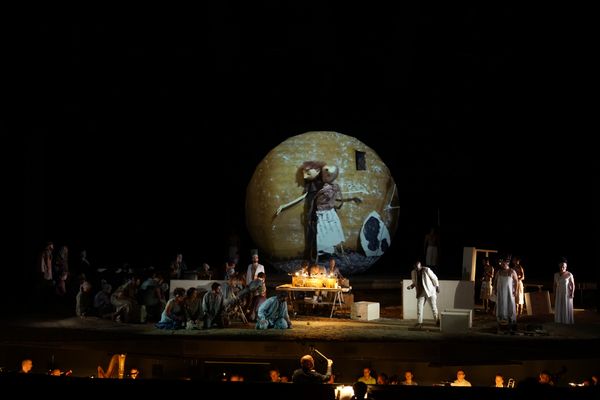 Wijd beeld van de opera met de grote bol waarop een figuur met een staf is geprojecteerd, het koor eromheen en het orkest op de voorgrond.
