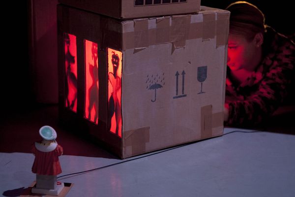 Een speelster buigt zich naar de kartonnen doos met de rood verlichte silhouetten; ervoor staat een poppetje met een lampje.