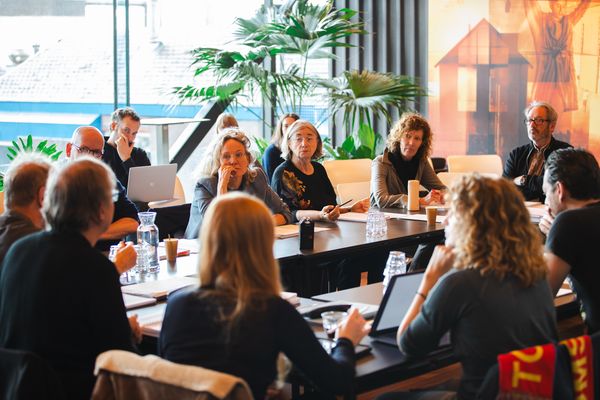 Een groep mensen zit rond samengeschoven tafels in een lichte zaal met palmen te overleggen.