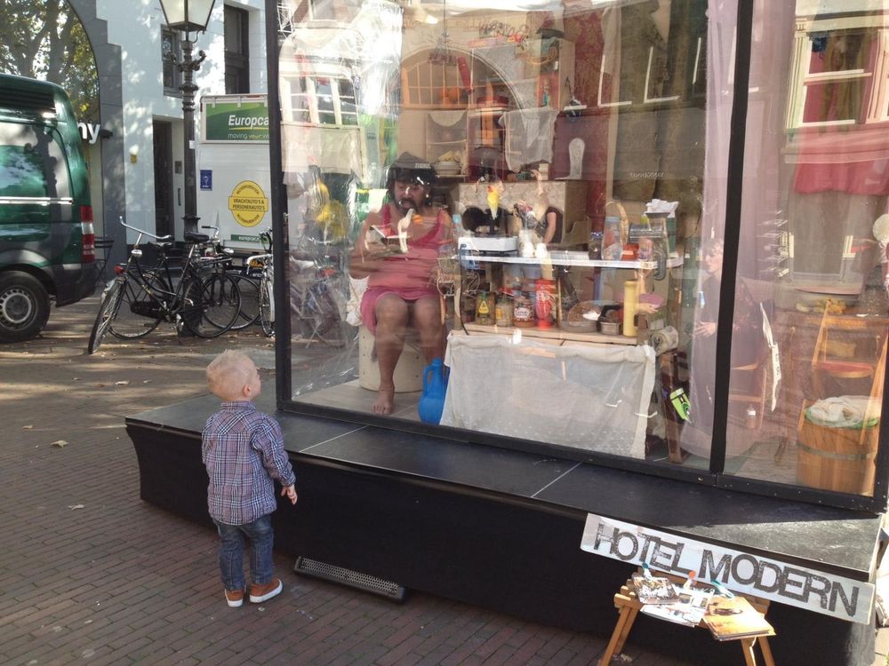 Een klein kind kijkt naar de glazen woonkubus op straat waarin een speelster met een hoed zit te eten, met fietsen en een bestelbus erachter.