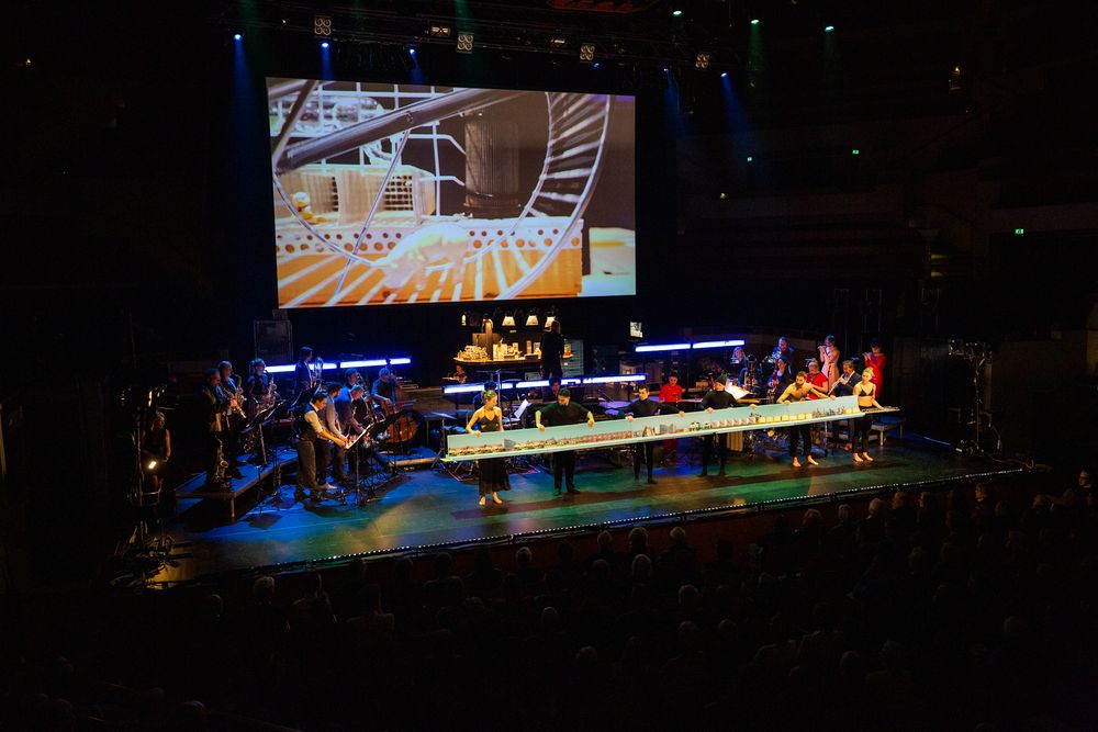 Concertzaal: op het scherm een gouden reuzenrad; op het podium spelen de blazers achter een lange tafel vol miniatuurdecors.