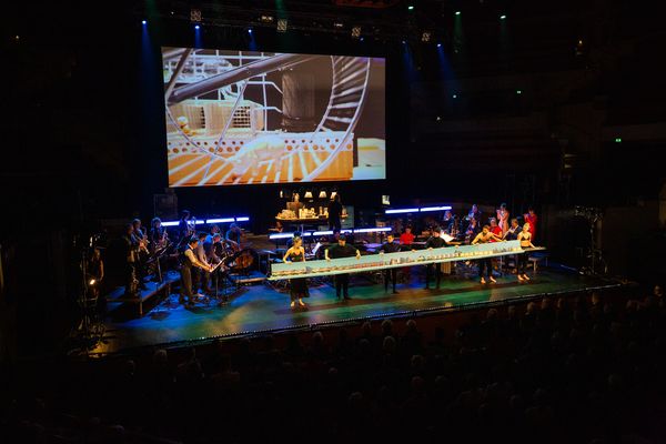 Concertzaal: op het scherm een gouden reuzenrad; op het podium spelen de blazers achter een lange tafel vol miniatuurdecors.