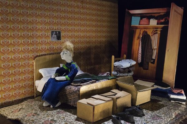 Miniatuurslaapkamer met bloemetjesbehang: een blonde pop zit op het bed tussen opgevouwen kleren, naast een open kledingkast en kartonnen dozen op het tapijt.
