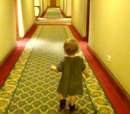 Videobeeld van een peuter in een groen jurkje die van de camera weg loopt door een lange hotelgang met gebloemd tapijt.