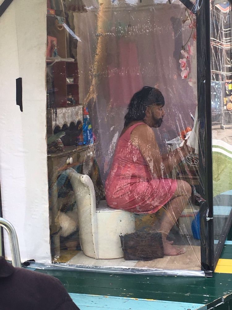 Een speler in een roze jurk met lang donker haar zit ineengedoken in een klein glazen huisje vol spullen.