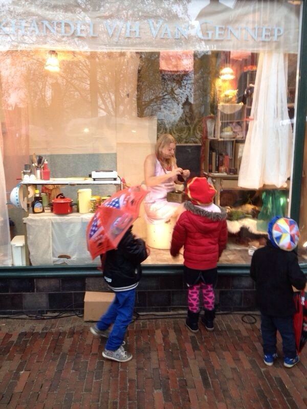 Een vrouw in een roze jurk zit te werken in de etalage van boekhandel Van Gennep; buiten kijken drie kinderen met paraplu's naar binnen.