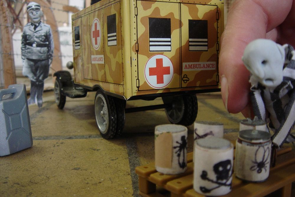 Een speelgoedambulance staat in de maquette; een hand zet er een poppetje bij, ernaast blikjes met een doodshoofd erop en een uitgeknipte foto van een bewaker.