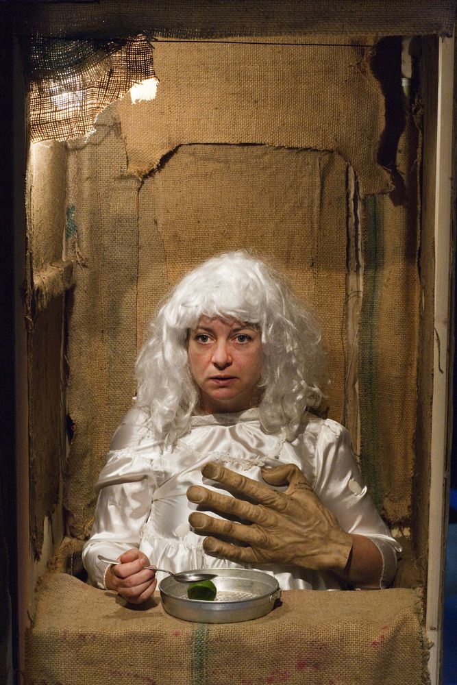 Een vrouw met een witte pruik zit in een met jute beklede kamer; over de tafel met een blikje schuift een reusachtige hand haar kant op.