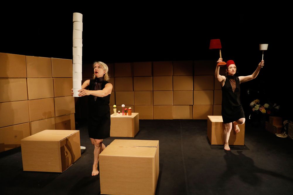 Arlène Hoornweg en Pauline Kalker in zwarte jurken op een podium van kartonnen dozen; de een houdt een torenhoge stapel wc-rollen omhoog, de ander balanceert een rode en een witte lampenkap.