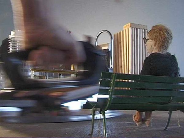 Een draaimolen tolt in beweging langs de camera; op een bankje ernaast zit een oude vrouw met haar rug naar ons toe.