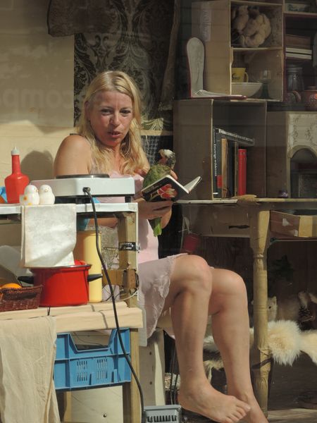 Een vrouw in een roze jurk leest met de benen over elkaar in haar etalagekamer, de papegaai op zijn stok naast haar.