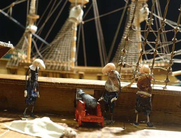 Drie poppen staan aan de reling van een miniatuurzeilschip naast een rood kanon, met masten en touwladders erachter.