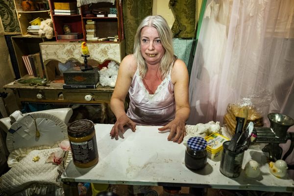 Een speelster in een roze hemdjurk staat grijnzend achter haar keukentafel vol potten en blikken in een volgestouwde kamer.