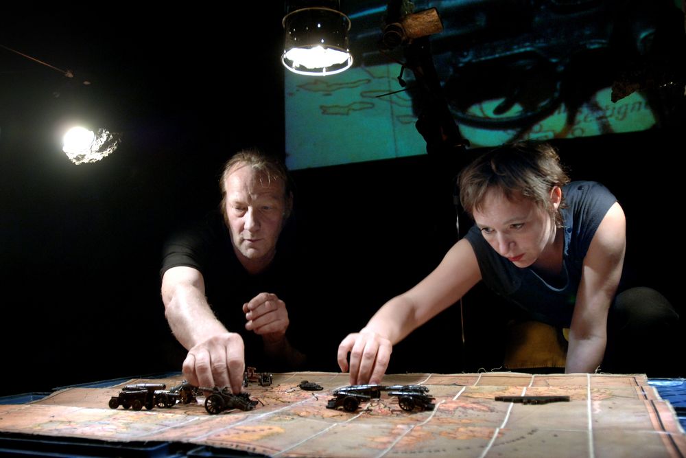 Twee spelers buigen zich over een tafel met een landkaart en zetten daar kleine kanonnen en voertuigen op; boven hen hangen werklampen.