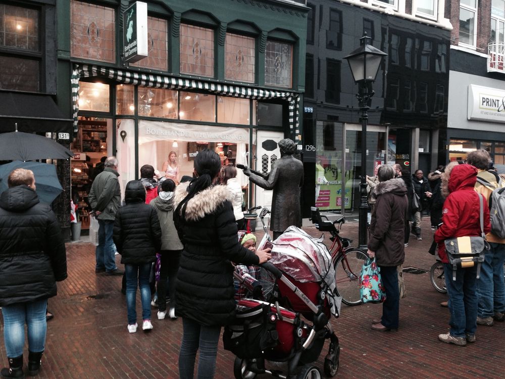 Voorbijgangers met kinderwagens blijven staan bij de etalage van de boekhandel; ernaast staat een standbeeld op de stoep.