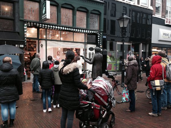 Voorbijgangers met kinderwagens blijven staan bij de etalage van de boekhandel; ernaast staat een standbeeld op de stoep.