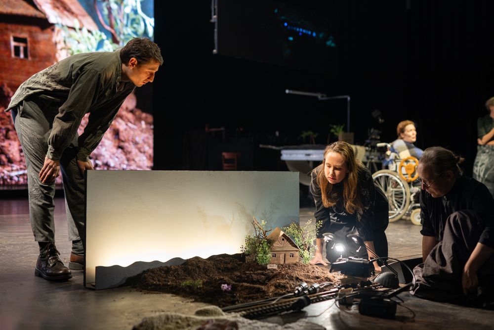 Een speler buigt zich over een miniatuurlandschap met een huisje op de toneelvloer, terwijl twee anderen het met een lamp en camera filmen.