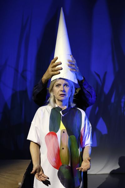 Twee handen zetten een hoge witte punthoed op het hoofd van een speelster in een beschilderd wit hemd, tegen een blauw doek met rietschaduwen.