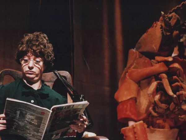 Een vrouw met een bril leest een tijdschrift in een fauteuil, naast haar torent een gestalte van schaaldierschalen op.