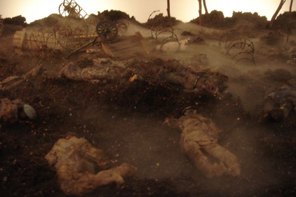 Miniatuurslagveld in sepia: gevallen soldaatjes liggen in de omgewoelde aarde tussen prikkeldraad, een omgevallen kar en kale boomstronken in de mist.